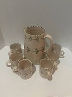 Full view of the ceramic pitcher surrounded by six matching coffee mugs, showing blush sand color with green vine pattern and scalloped rim on pitcher.