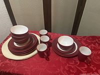 Overview photo of entire 16 pc set showing plates, bowls, cups stacked and four placemats underneath on red surface.