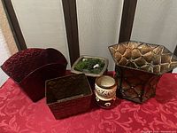 Photo showing a red ceramic semi-circular pot, dark brown square pot, square stone-like planter with moss, small beige and brown ceramic pot with cut-outs, and a large decorative metal pot with gold tones on a patterned red tablecloth.