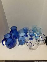 Full set of 7 blue glassware items including two cobalt blue pitchers, one acrylic blue pitcher, two small drinking glasses, and two margarita glasses with swirl pattern.