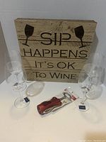 Four clear Mikaka wine glasses, four Royal Leerdam champagne glasses, a wooden sign reading 'SIP HAPPENS IT'S OK TO WINE', and a red wine opener packaged, on white surface.
