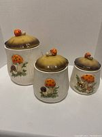 Three ceramic mushroom canisters of different sizes with lids, featuring textured bodies and mushroom decorations on the front and tops.