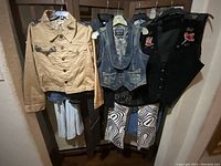 Photo showing four hanging garments: beige jean jacket with butterfly patches, blue denim lace vest, black floral embroidered vest, and abstract pattern pants.
