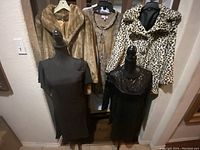 Six pieces of ladies clothing on hangers and mannequins: four dresses and two faux fur coats in a hallway area.