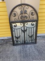 View of entire wrought iron arched garden gate mirror showing decorative scrollwork and butterfly details with reflective mirrors