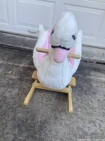 Front angled view of the plush swan rocker showing white and pink plush body, wooden handles and base on outdoor concrete surface.
