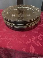 Stack of nine round plate chargers with gold embossed floral and geometric patterns on red base.