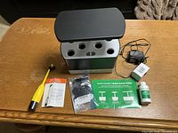 AeroGarden unit top view showing six plant pods, power adapter plug, Bee Pollinator wand, replacement filters, plant food bottle, and printed instructions on a wooden table.