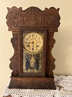 Front view of antique wooden gingerbread clock with ornate carvings and glass door showing clock face and pendulum.
