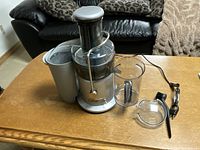 Breville electric juicer with waste bucket on the left, juice jug, juice jug lid, cleaning brush, and plunger on a wood table with couch background