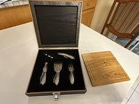 Setup of cheese board next to open black-lined storage case with cheese utensils and wine accessories inside, new condition, case top damaged.