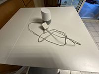 White Google Home smart speaker with power adapter and cable on a white table, photographed from the side and front angles.
