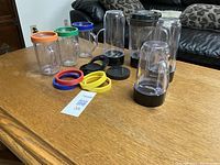 Photo of multiple clear plastic blender cups with handles, several colorful sealing rings, black lids, and a 4-blade assembly base on a wooden table with black leather couch in the background.