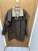 Front view of Koolah brown oilskin coat hanging on hanger showing 3/4 length, tan collar, and detachable cape along the back.