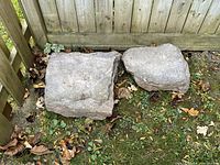 Two large natural gray landscaping rocks side by side on grass near a wooden fence, showing size and texture.