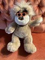Front view of the Bubba bear plush toy seated on an orange chair with its right arm raised, wearing bright orange sunglasses over dark patch eyes and white fluffy hair.