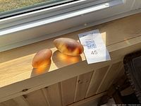 Two oval-shaped amber to brown colored sea glass pieces sitting on a wooden windowsill with strong natural light.