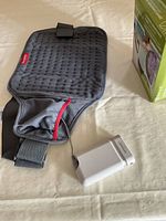 Sunbeam gray quilted rechargeable heating pad with remote and charging adapter visible on a beige surface with part of the product box shown.