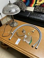 Photo showing all items on a wooden coffee table including the decorated glass shade table lamp, two washer hoses, circular metal hand towel rack with screws, three door knobs with hardware, and a plastic bag of mounting screws.