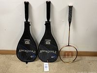 Two Wilson Pro Staff tennis racquets with black covers and one badminton racquet with reddish-brown frame and white grip.