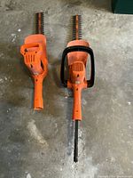 Pair of Black & Decker electric hedge trimmers shown side by side with 16-inch blades and orange bodies on a concrete floor.