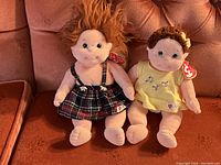 Front view of both Ty Beanie Kids dolls seated on a cushioned sofa showing Curly on left with plaid jumper and Ginger on right in yellow dress with tags attached.