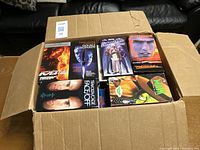 Open cardboard box showing several VHS tapes packed inside. Visible titles include Double Jeopardy, The Devil's Advocate, My Best Friend's Wedding, Face/Off, The Mask, and more.