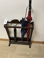 Front view of vintage dark-stained wooden umbrella stand holding three umbrellas, black, navy, and red-striped, showing metal drip tray at base.