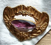 Close-up front image of the antique Art Deco style brooch showing the large faceted amethyst purple-colored cut-glass centerpiece set in the ornate embossed bronze frame with floral motifs.