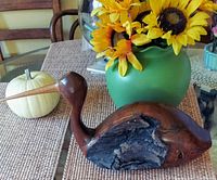 Side view of wooden shore-bird sculpture with natural wood grain and burl textures, placed next to a green vase with sunflowers and decorative white pumpkin on woven fabric surface.