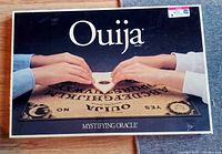 Photo of the Ouija board game in its box showing two hands using the planchette and the board details