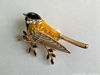 Front view of yellow bird brooch showing black head, yellow enamel body, and clear rhinestone neck band, perched on gold-tone branch with green leaves.