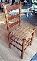 Side angle view of the old Shaker side chair showing its wooden frame and woven rush seat on hardwood floor with surrounding furniture.