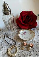 Clear ribbed glass talc shaker with silver-plated lid, small Haviland porcelain dish, large red silk rose, pearl enamel gold-tone earrings with rhinestones and pearl drops, and pearl and crystal pendant necklace on black cord, all displayed on a white lace fabric background.