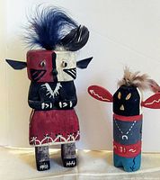Front view of two carved wooden Native American Kachina figures; one larger with red, black, and white colors and feather on head, the other smaller with black, red, blue colors and feather on top.