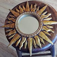 Full view of vintage sunburst wall mirror on round wooden stool showing gilded carved frame with alternating straight and wavy rays and central round mirror.