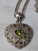 Close-up image of the heart-shaped silver pendant showing detailed filigree work and central green heart-shaped gemstone.
