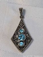 Front angled photo of sterling silver pendant with three blue round stones and marcasite accents on a white textured background.