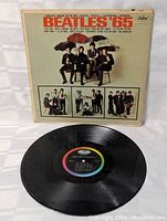 Front cover of 'The Beatles 65' vinyl LP with band members holding umbrellas, listing song titles at the top.
