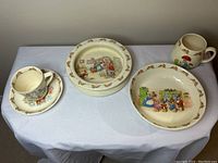 Overhead view of six Bunnykins ceramic items on white cloth showing mugs, bowls, plates with bunny illustrations and borders.