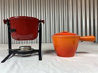 Full view of red fondue stand on left and orange pot with lid on right