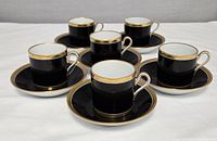 Six black porcelain espresso cups with white interior and gold trim each placed on matching saucers with gold rims.