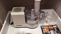 Full view of Braun food processor with motor base, clear processing bowl with handle, feed tube, accessories, and cookbook on white surface