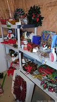 Wide angle showing various Christmas decorations including snowman figurine, wreaths, jars, ribbons, mugs, and gift bags arranged on white shelving units.
