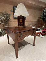 Wood end table with dark finish and three front drawers. A table lamp with decorative cylindrical base and white pleated lampshade sits on top along with three wooden coasters.