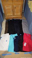 Full view of eight pairs of golf pants in various colors and one red golf shirt with tag, displayed on floor in two rows, with wooden cabinet backdrop.