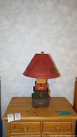 Wood lamp on wooden dresser with light blue and gray cloud patterned wall behind. The red metal shade shows punched decorative patterns, the base shows block design with year '1791' and a small drawer.
