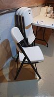 Four white plastic folding chairs with dark metal frames stacked against a wall in a basement setting.