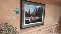 Framed limited edition print hanging on a beige wall above white wainscoting. Shows voyageurs in canoes on a river with a mountainous forest background in natural colors. The frame is dark wood with double green and white mattes.