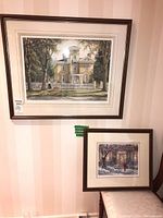 Both framed prints displayed on a wall with a striped wallpaper background, showing the larger yellow house scene and the smaller winter scene with a woman and child.